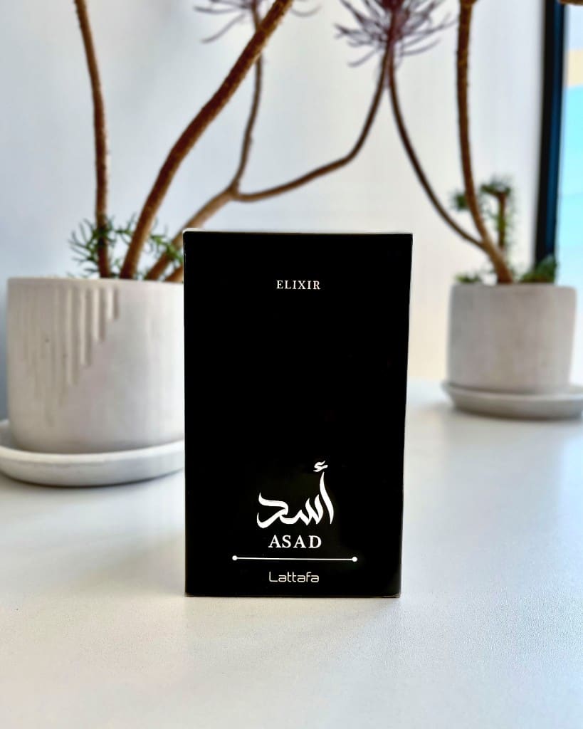 Caja de Asad Elixir de Lattafa sobre una mesa con plantas de fondo